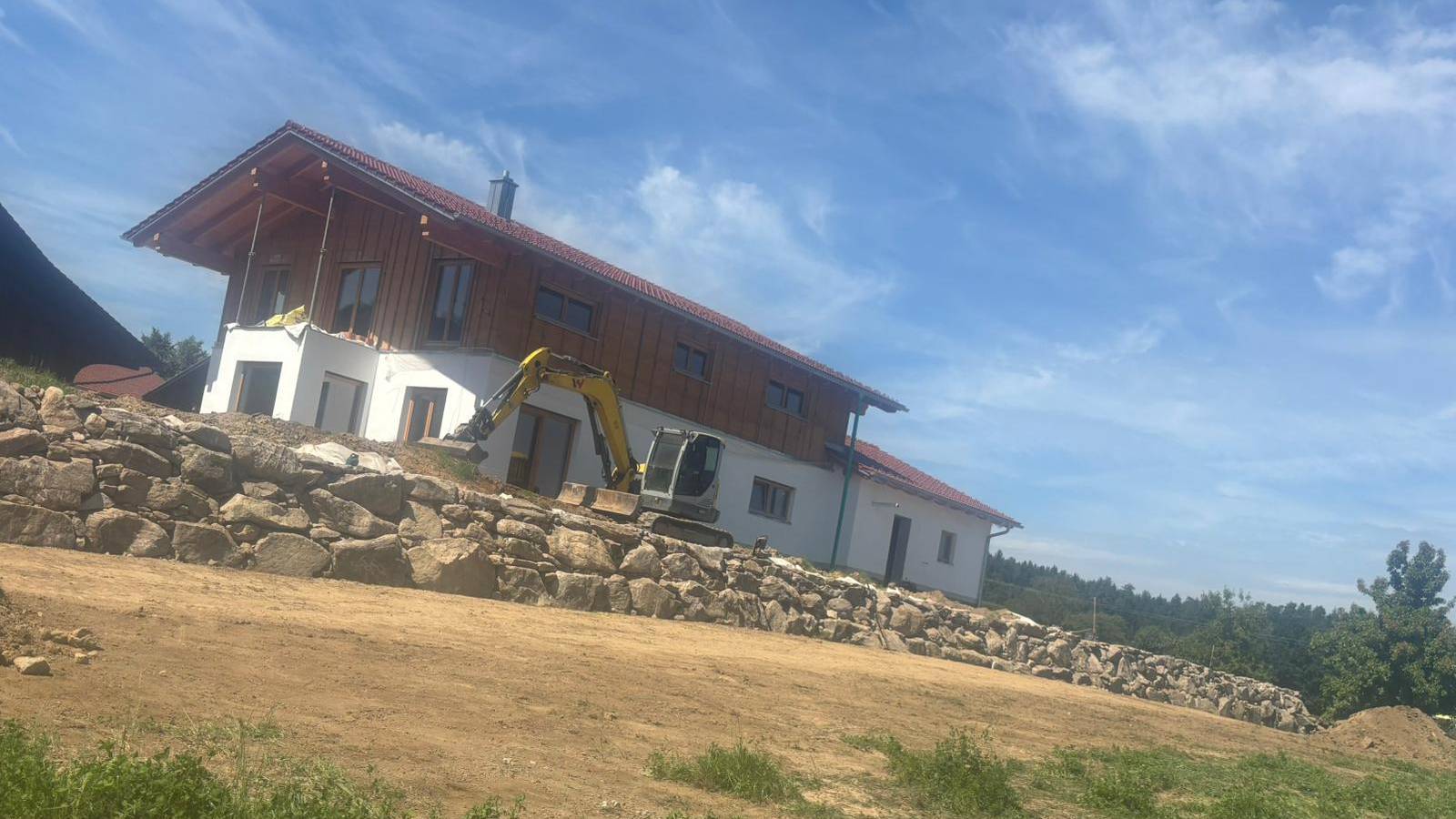 Kettenbagger, Natursteinmauer aus Granit-Findlingen und fertiges Wohnhaus – Außenanlage von Forst- & Baggerbetrieb Kronfeldner im Bayerischen Wald